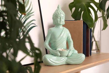 Ceramic Buddha sculpture with burning candle and books on wooden shelf indoors. Interior decorの写真素材