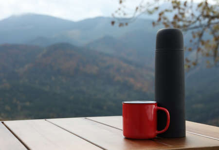 Black thermos and metal mug on wooden table against mountain landscape. Space for textの写真素材