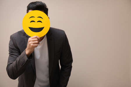 Man hiding emotions using card with drawn smiling face on beige background. Space for textの写真素材