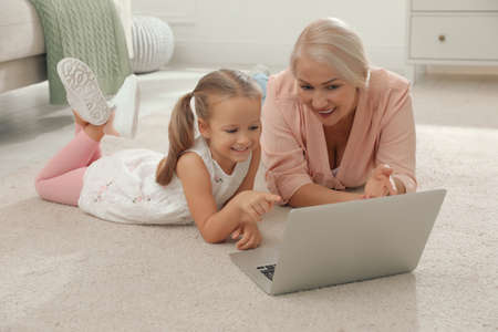 Happy grandmother and her granddaughter using laptop together on floor at homeの写真素材