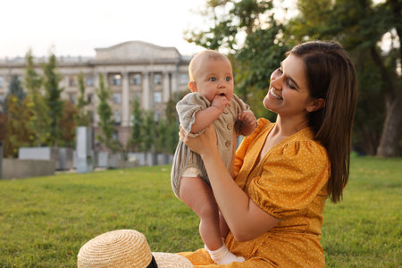 Happy mother with adorable baby sitting on green grass in park, space for text