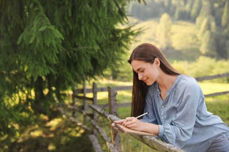 Beautiful young woman drawing with pencil in notepad near wooden fenceの写真素材