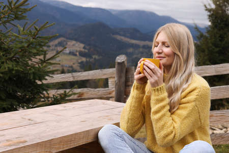 Young woman with mug of hot drink at wooden table in mountainsの写真素材