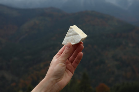 Woman holding piece of delicious cheese against mountain landscape, closeup
