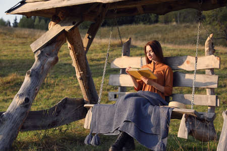 Beautiful young woman reading book on wooden swing outdoorsの写真素材