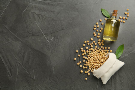 Glass bottle of oil, leaves and soybeans on gray table, flat lay. Space for text