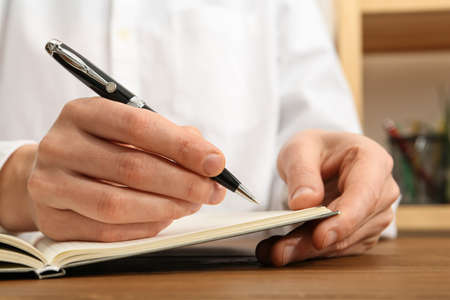 Man writing with pen in notebook at wooden table, closeup of hands