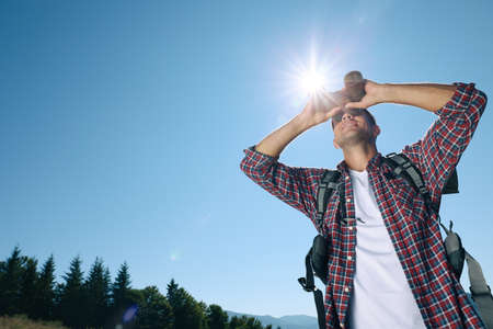 Tourist with hiking equipment looking through binoculars outdoors on sunny day, low angle viewの写真素材