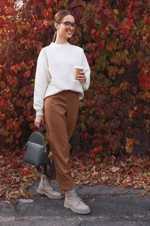 Beautiful young woman with stylish gray backpack and hot drink in autumn parkの写真素材