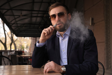 Handsome young businessman using disposable electronic cigarette at table in outdoor cafeの写真素材