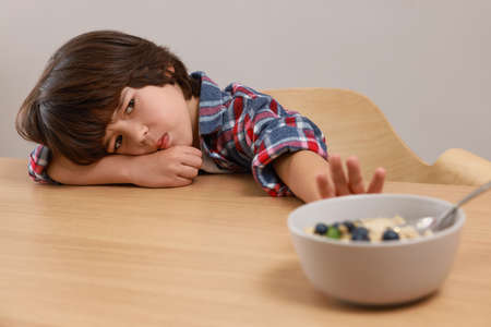 Cute little boy refusing to eat his breakfast at table on gray backgroundの写真素材