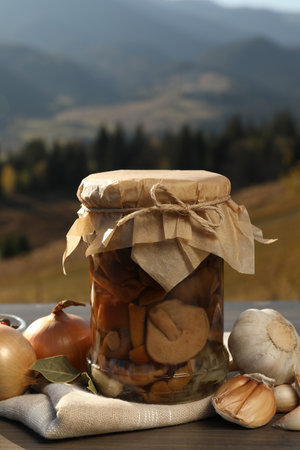 Glass jar with pickled mushrooms and fresh ingredients on wooden table against mountain landscape. Space for textの写真素材