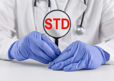 Doctor holding magnifying glass with abbreviation STD at white table, closeupの写真素材