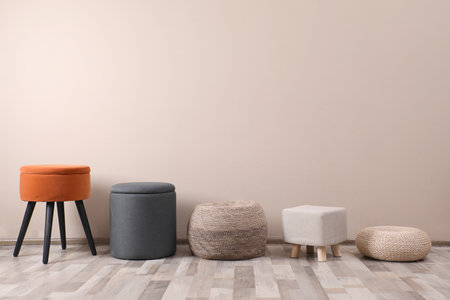 Different stylish poufs and ottomans near beige wall, space for text