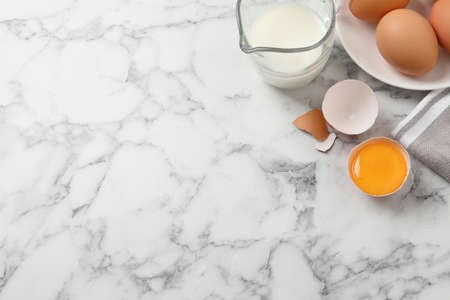 Raw chicken eggs, shell with yolk and jug of milk on white marble table, flat lay. Space for textの写真素材