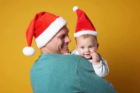 Happy father with cute baby wearing Santa hats on yellow background. Christmas seasonの写真素材