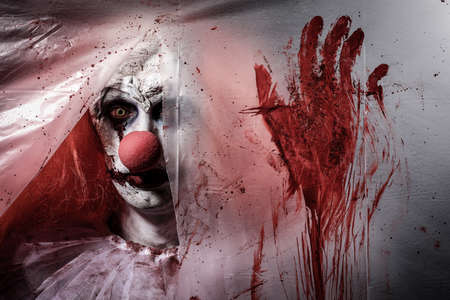 Terrifying clown staring through hole in torn bloodstained plastic film. Halloween party costumeの写真素材