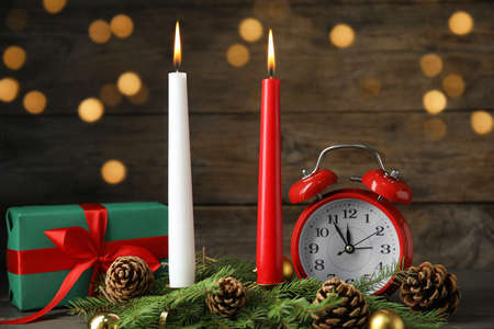 Burning candles, festive decor, gift box and alarm clock on wooden table against blurred Christmas lightsの写真素材