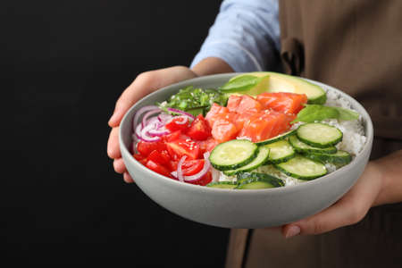 Woman holding delicious poke bowl with salmon and vegetables on black background, closeupの写真素材