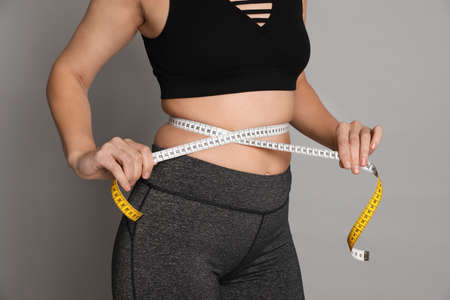 Woman measuring waist with tape on gray background, closeup