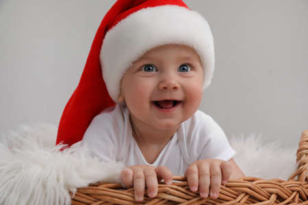 Cute baby wearing Santa hat in wicker basket on light gray background. Christmas celebration