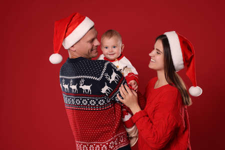 Happy couple with cute baby in Christmas outfits and Santa hats on red backgroundの写真素材