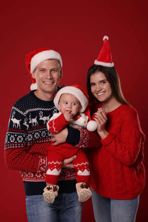 Happy couple with cute baby in Christmas outfits and Santa hats on red backgroundの写真素材