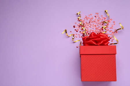 Red gift box with sequins and streamers on lilac background, flat lay. Space for textの写真素材