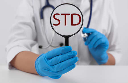 Doctor holding magnifying glass with abbreviation STD at white table, closeupの写真素材