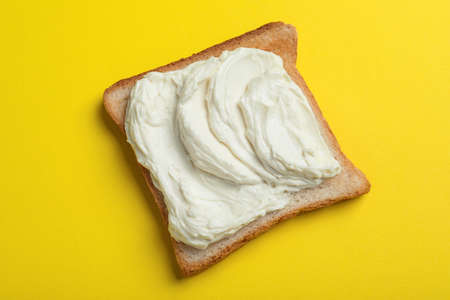 Slice of bread with tasty cream cheese on yellow background, top view