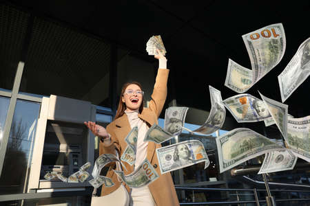 Excited young woman with money near cash machine outdoorsの写真素材