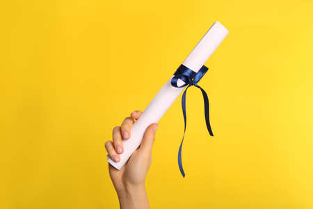 Student holding rolled diploma with blue ribbon on yellow background, closeupの写真素材
