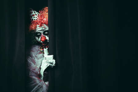 Terrifying clown hiding behind black curtains, space for text. Halloween party costume