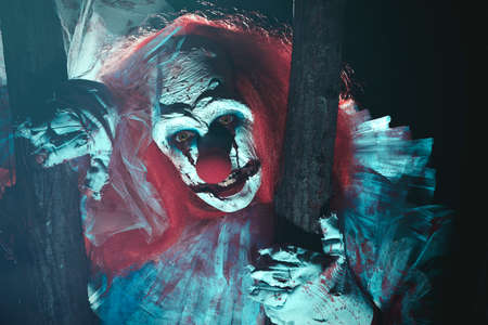 Terrifying clown hiding behind trees outdoors at night. Halloween party costume