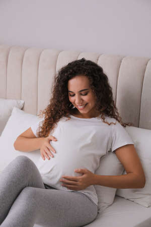 Pregnant young African-American woman sitting on bed at homeの写真素材
