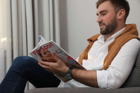 Handsome man reading magazine in armchair indoors
