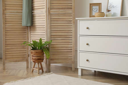 Stylish room with wooden folding screen and chest of drawers near white wall. Interior design