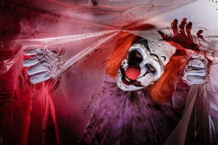 Terrifying clown staring through hole in torn bloodstained plastic film. Halloween party costumeの写真素材