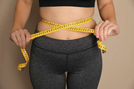 Woman measuring waist with tape on beige background, closeup