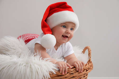 Cute baby wearing Santa hat in wicker basket on light gray background. Christmas celebrationの写真素材
