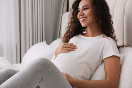 Pregnant young African-American woman sitting on bed at homeの写真素材
