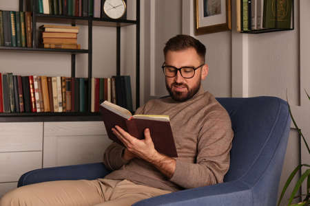 Young man reading book in armchair indoors. Home library