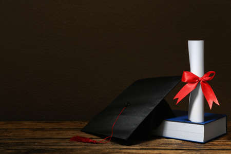 Graduation hat, book and diploma on wooden table against brown background, space for textの写真素材
