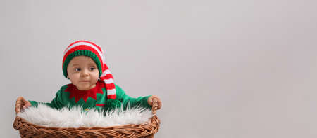 Cute baby wearing elf costume near wicker basket on light gray background, banner design with space...