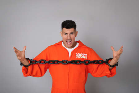 Emotional prisoner in orange jumpsuit with chained hands on gray background