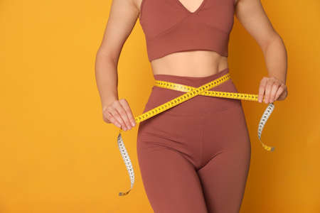 Woman in sportswear measuring waist with tape on yellow background, closeup. Space for textの写真素材