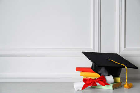 Graduation hat, books and diploma on floor near white wall, space for textの写真素材
