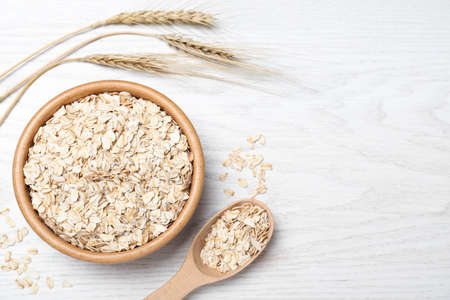 Oatmeal, bowl, spoon and spikelets on white wooden table, flat lay. Space for text