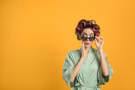 Emotional young woman in silk bathrobe with hair curlers and sunglasses on yellow background. Space for textの写真素材