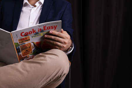 Man reading magazine in armchair against dark background, closeup. Space for text